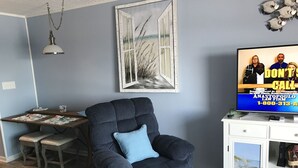 Olohuoneessa on tummansininen nojatuoli, valkoinen kaappi, josta avautuu näkymä rannalle, ja televisio, jossa näkyy lakitoimiston mainos.