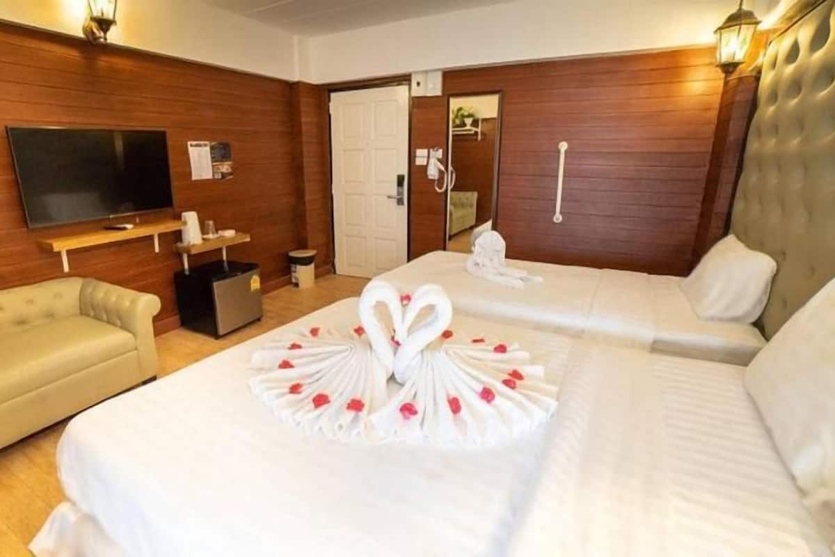 A hotel room with a wooden wall, a television, a sofa, two beds with white bedding and a heart-shaped towel arrangement, and a small trash bin.