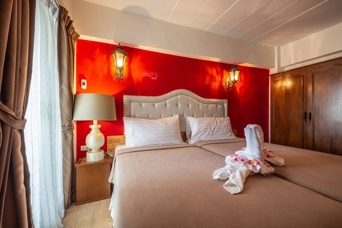 A hotel room with a red wall, a white bed with a tufted headboard, two bedside lamps, a wooden nightstand, and a wooden wardrobe.