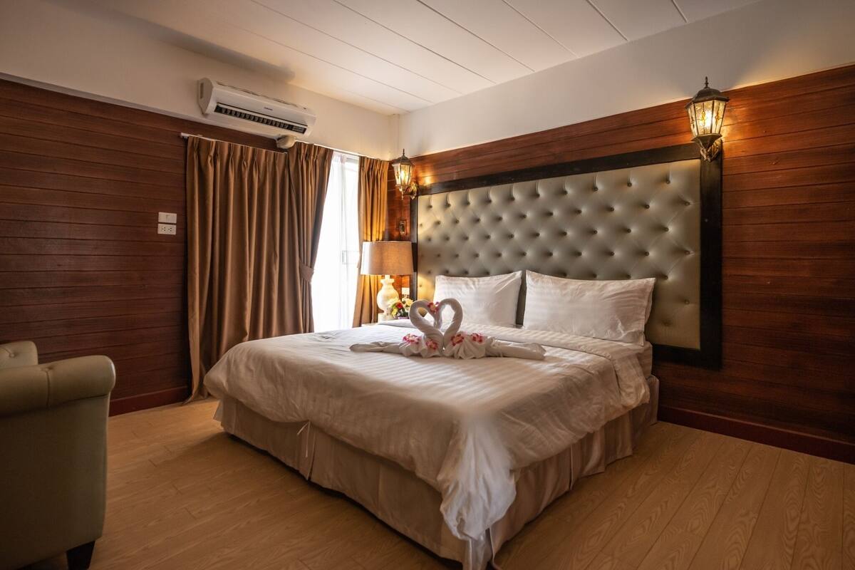 A hotel room with a large bed, wooden panel walls, a tufted headboard, and a nightstand with a lamp.
