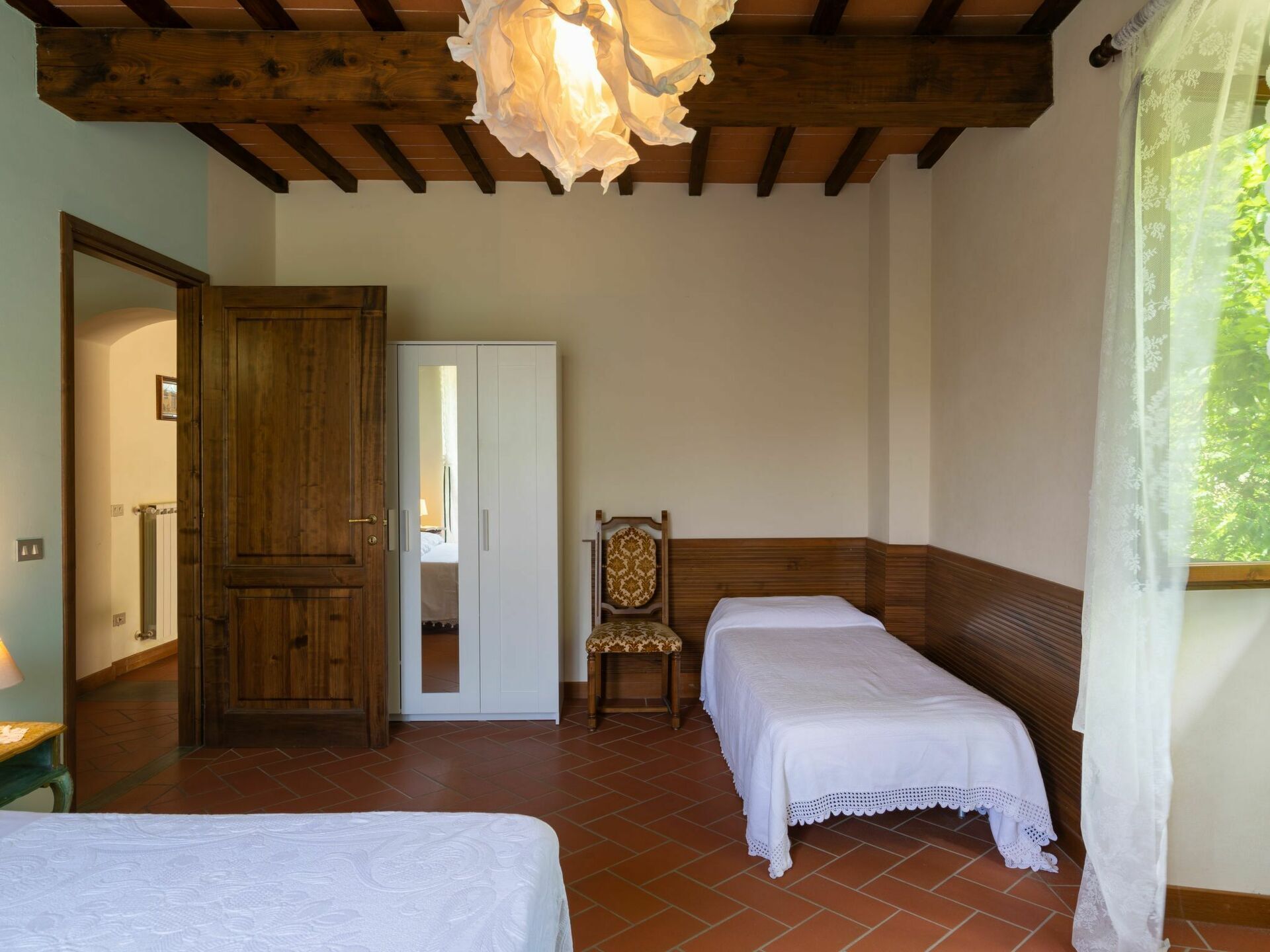 Une chambre avec deux lits, une armoire en bois, une chaise et une fenêtre donnant sur un paysage verdoyant.