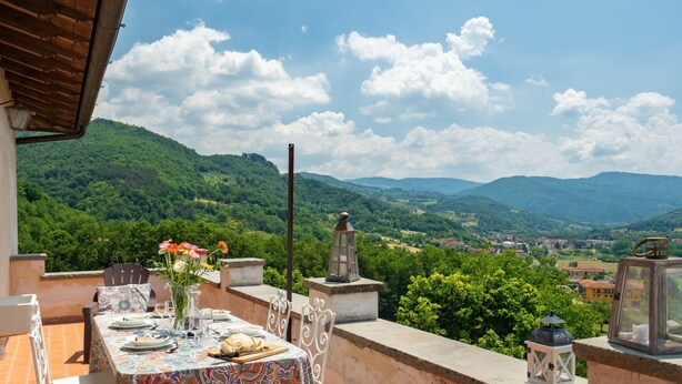Terraço com mesa posta para refeições, com vista para uma paisagem montanhosa.