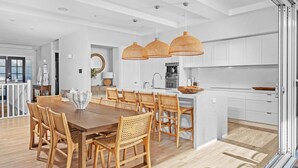 Una cocina moderna con armarios blancos, una isla central y un comedor con mesa de madera y sillas de mimbre.
