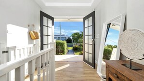 Pasillo con barandillas blancas, suelo de madera y vistas a una casa y jardín a través de puertas abiertas.