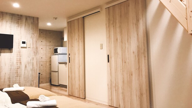 Ein modernes Hotelzimmer mit Holzboden, einem Bett mit Handtüchern, einer kleinen Küchenzeile und einer schiebbaren Holztür.