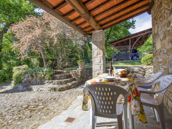 Un patio al aire libre con suelo de piedra, una mesa dispuesta con una botella de vino, pan y fruta, y dos sillas de plástico.