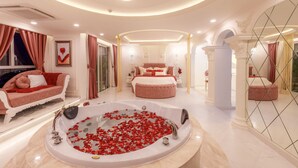 A luxurious bedroom with a large bed, a sitting area with a sofa, a bathtub with rose petals, and a mirror wall.