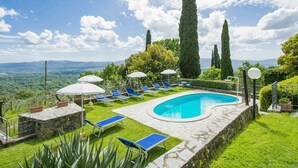 A swimming pool with lounge chairs and umbrellas, surrounded by greenery and tall trees, with a view of distant mountains. - Lovely apartment in villa for 4 people with pool, WIFI (Loro Ciuffenna)