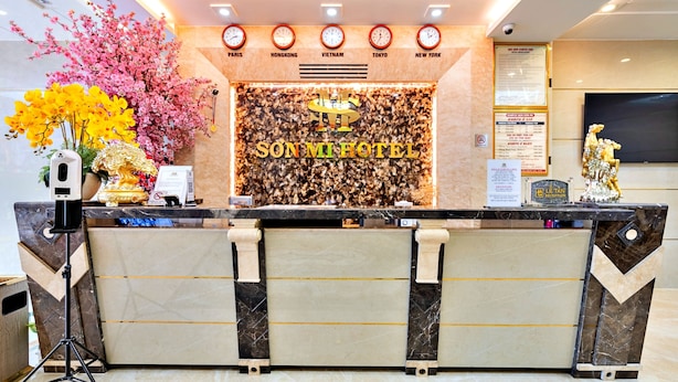 A hotel reception desk with a modern design, featuring a marble counter, a decorative floral arrangement, and a digital clock display. - S79 Son Mi Hotel 839 Le Hong Phong Street (Ho Chi Minh City)