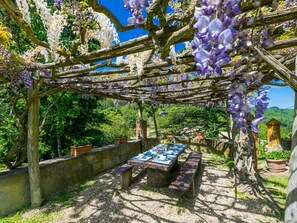 An outdoor dining area with a pergola covered in wisteria vines, surrounded by greenery and a stone wall. - Cosy apartment in villa for 10 guests with private pool, WIFI, TV, terrace and pets allowed (Greve In Chianti)