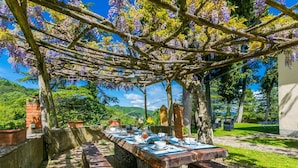 An outdoor dining area with a table set for two, surrounded by a pergola with purple flowers, a brick wall, and a view of greenery and hills. - Beautiful apartment in villa for 10 guests with private pool, WIFI, TV, terrace and pets allowed (Greve In Chianti)