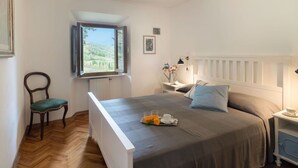 Een slaapkamer met een bed, een stoel, een nachtkastje, een lamp en een raam met uitzicht op het landschap.