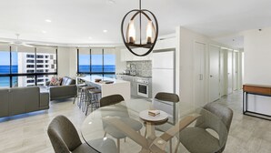 A modern living room with a glass dining table, grey sofas, and a kitchen area with a view of the ocean. - Victoria Square Private Apartments (Broadbeach)