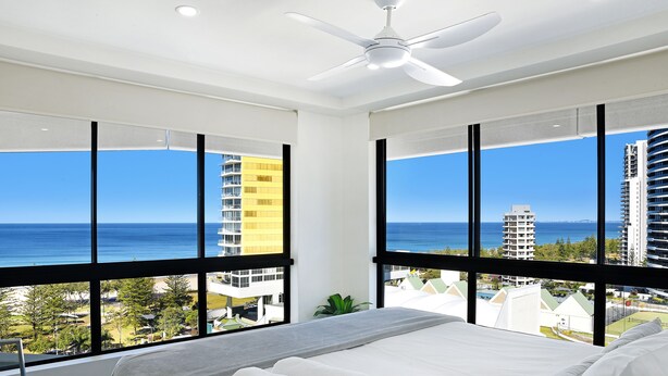 A hotel room with a large bed, two windows with ocean views, a ceiling fan, and a view of nearby buildings. - Victoria Square Private Apartments (Broadbeach)