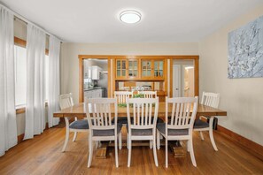 A dining area with a wooden table and white chairs, a kitchen in the background, and a painting on the wall. - Charming & Spacious Craftsman House! (Spokane)