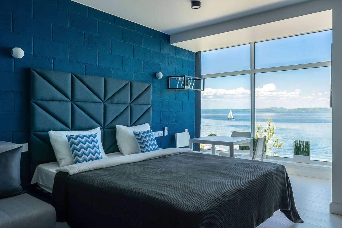 A modern bedroom with a large bed, a view of the sea, and a blue brick wall.