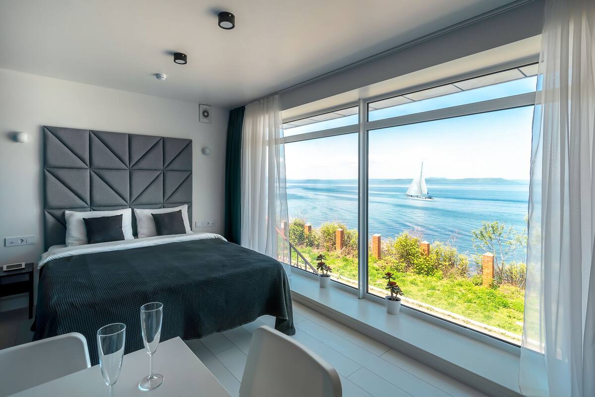 A modern bedroom with a large window, a bed, and a dining area with a view of the sea.