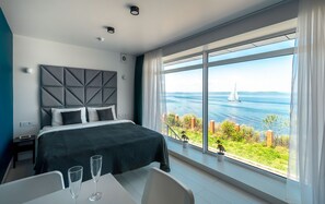 A modern bedroom with a large window, a bed, and a dining area with a view of the sea. - Panorama Sea View (Vladivostok)
