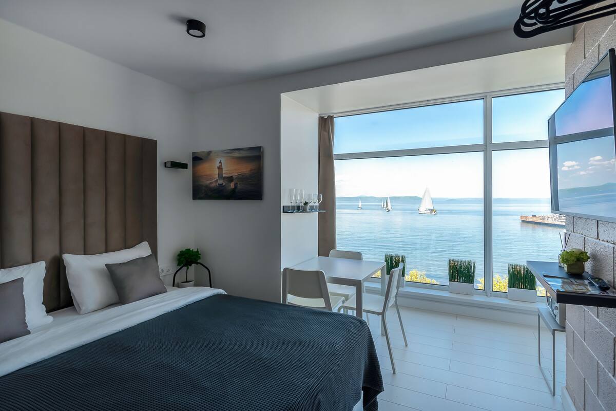 A modern hotel room with a large bed, a desk, and a balcony with a view of the sea.