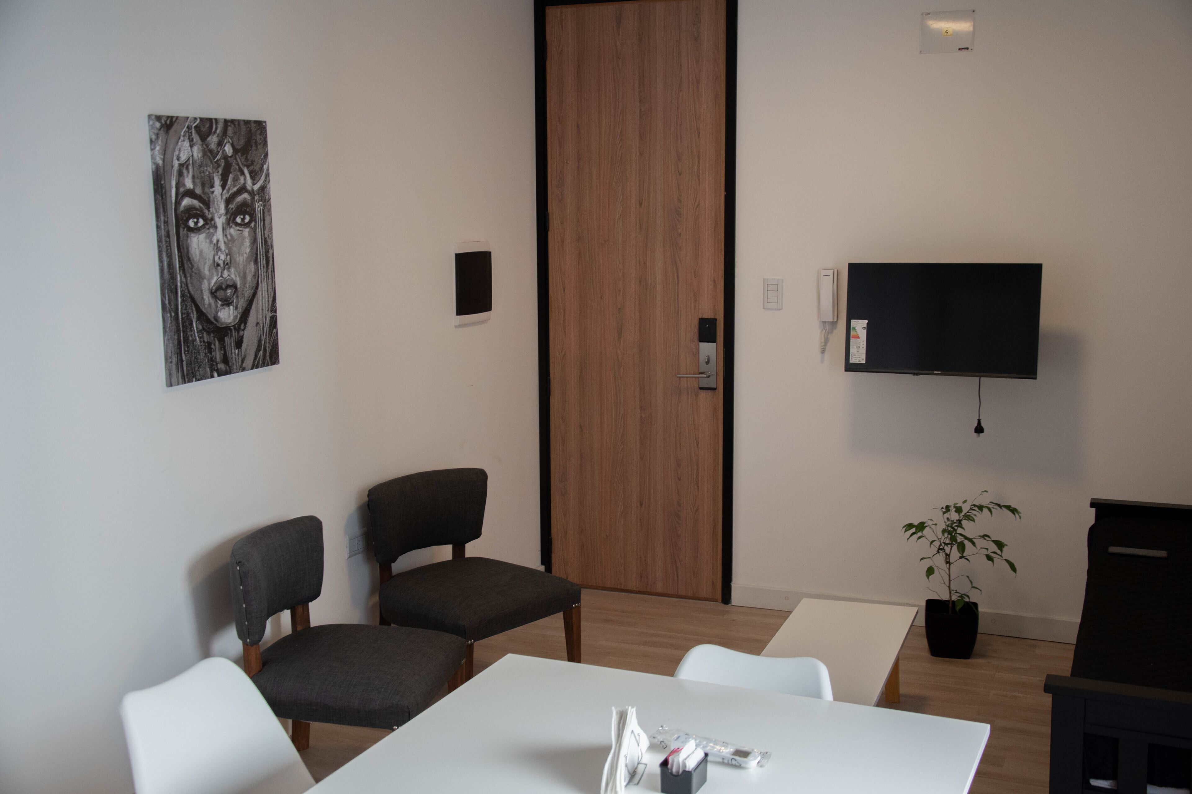 Una habitación moderna con una puerta de madera, un televisor de pantalla plana, dos sillones grises tapizados, una mesa blanca y una planta en maceta.