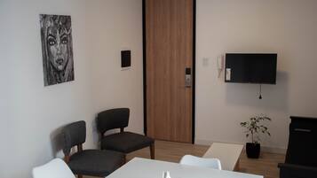 Una habitación moderna con una puerta de madera, un televisor de pantalla plana, dos sillones grises tapizados, una mesa blanca y una planta en maceta.