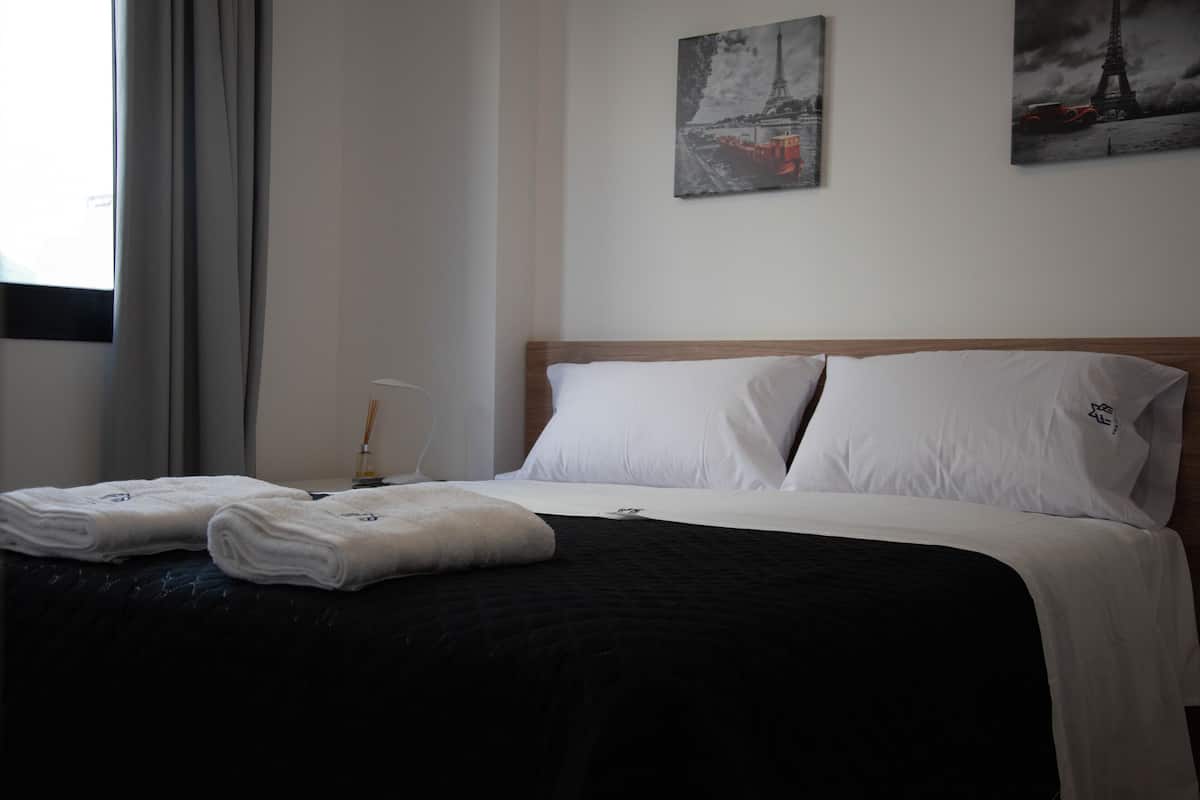A neatly made bed with white linens and a black bedspread, two white pillows, and a window with curtains.