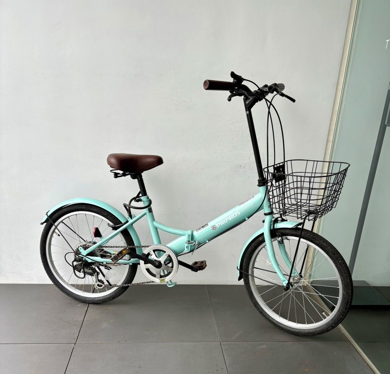 A teal bicycle with a brown seat, a front basket, and a rear rack, parked against a wall.