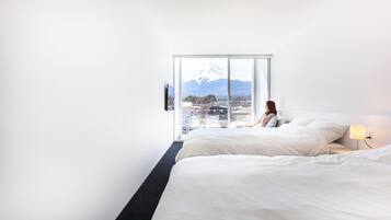 Ein Hotelzimmer mit großem Fenster, einem Bett, einem Nachttisch mit Lampe und Blick auf einen Berg.