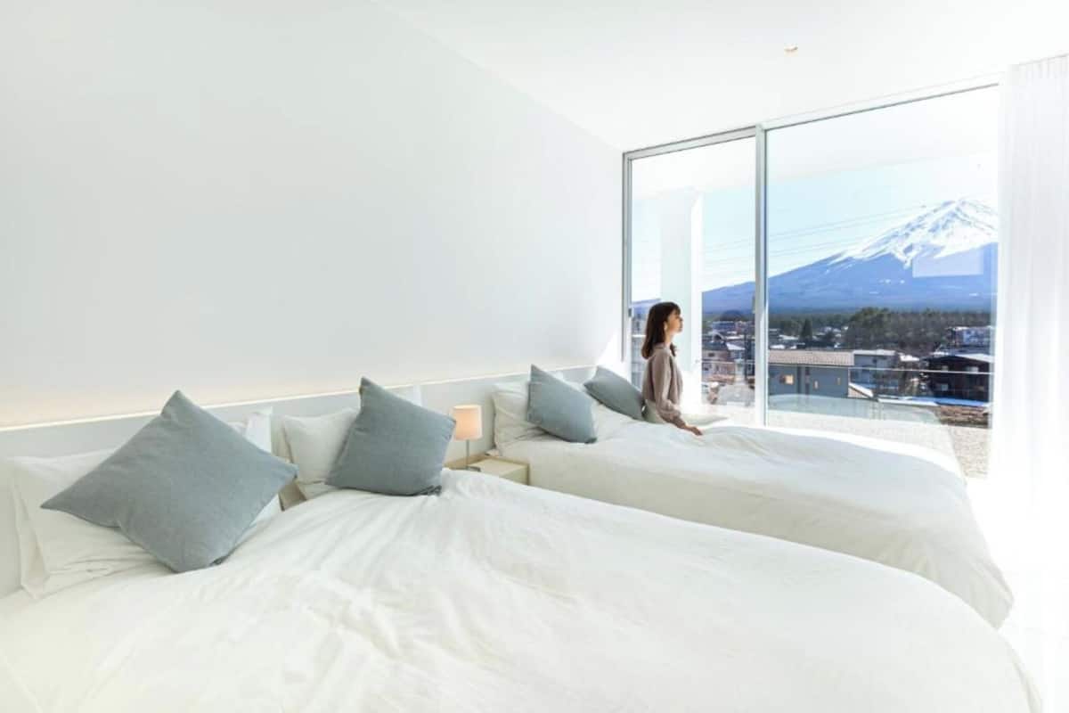 A modern hotel room with two beds, a large window with a mountain view, and a nightstand with a lamp.