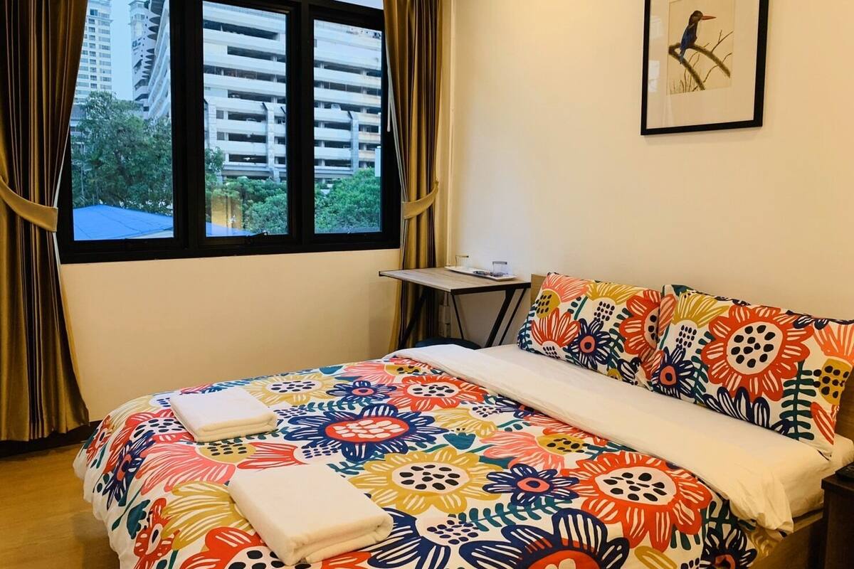 A hotel room with a bed, a small table, a chair, a window with curtains, and a framed picture on the wall.