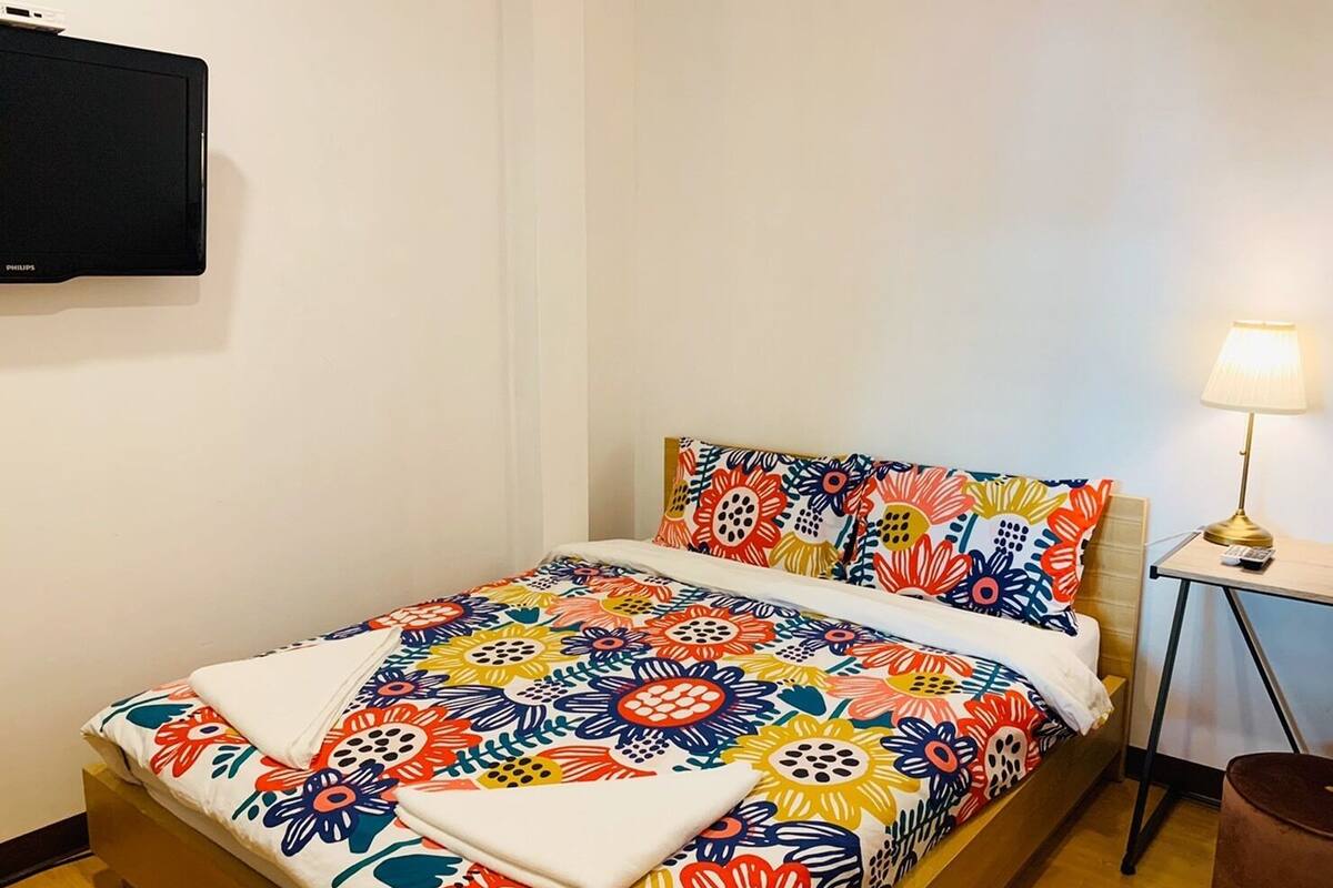 A hotel room with a bed, a TV mounted on the wall, a bedside lamp, and a small table with a lamp.