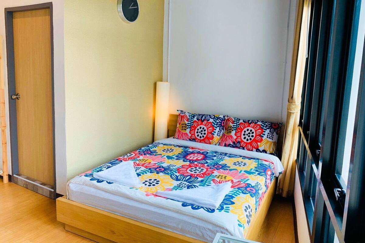A small, neatly arranged bedroom with a bed, a bedside lamp, a clock on the wall, and a window with blinds.