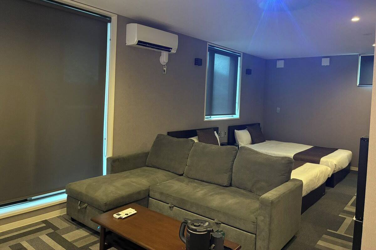 A hotel room with a sofa, two single beds, a TV, and an air conditioning unit.