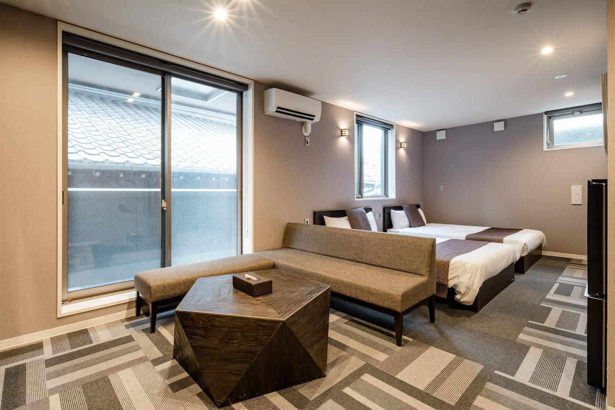 A hotel room with a large sliding glass door, a sofa bed, a small table, and a bed with two pillows.