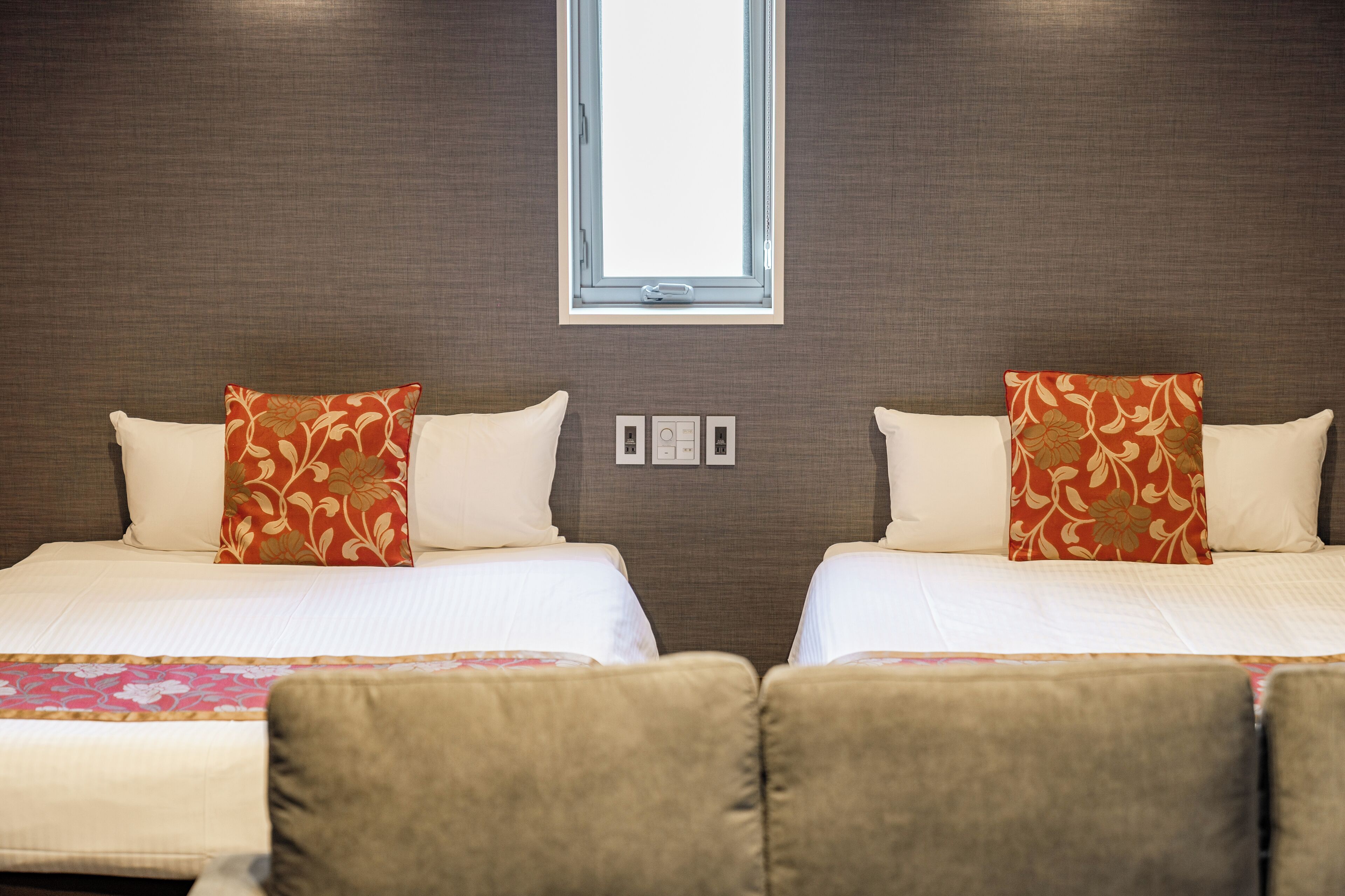Ein Hotelzimmer mit zwei Betten, jedes mit einem blumigen Kopfkissen, einem Fenster darüber und einer wandmontierten Steuerung.
