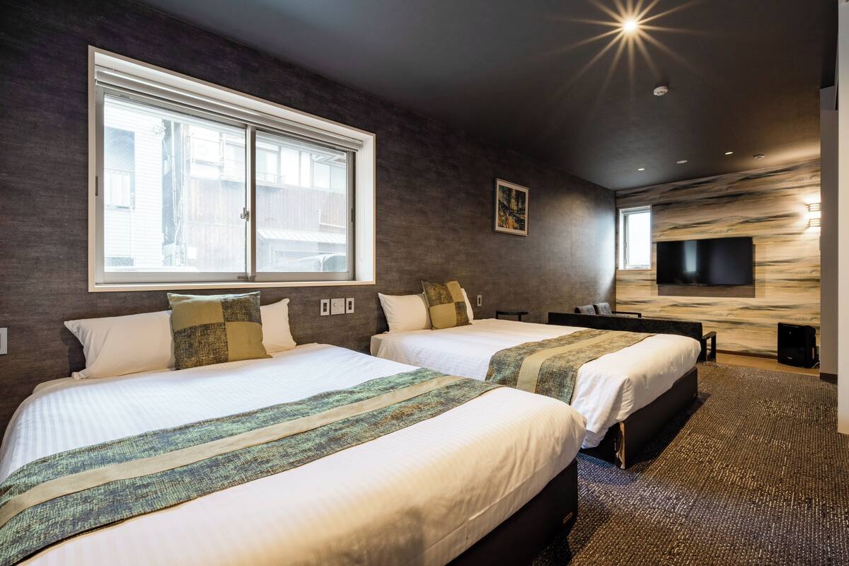 A hotel room with two beds, a large window, a flat-screen TV mounted on the wall, and a carpeted floor.