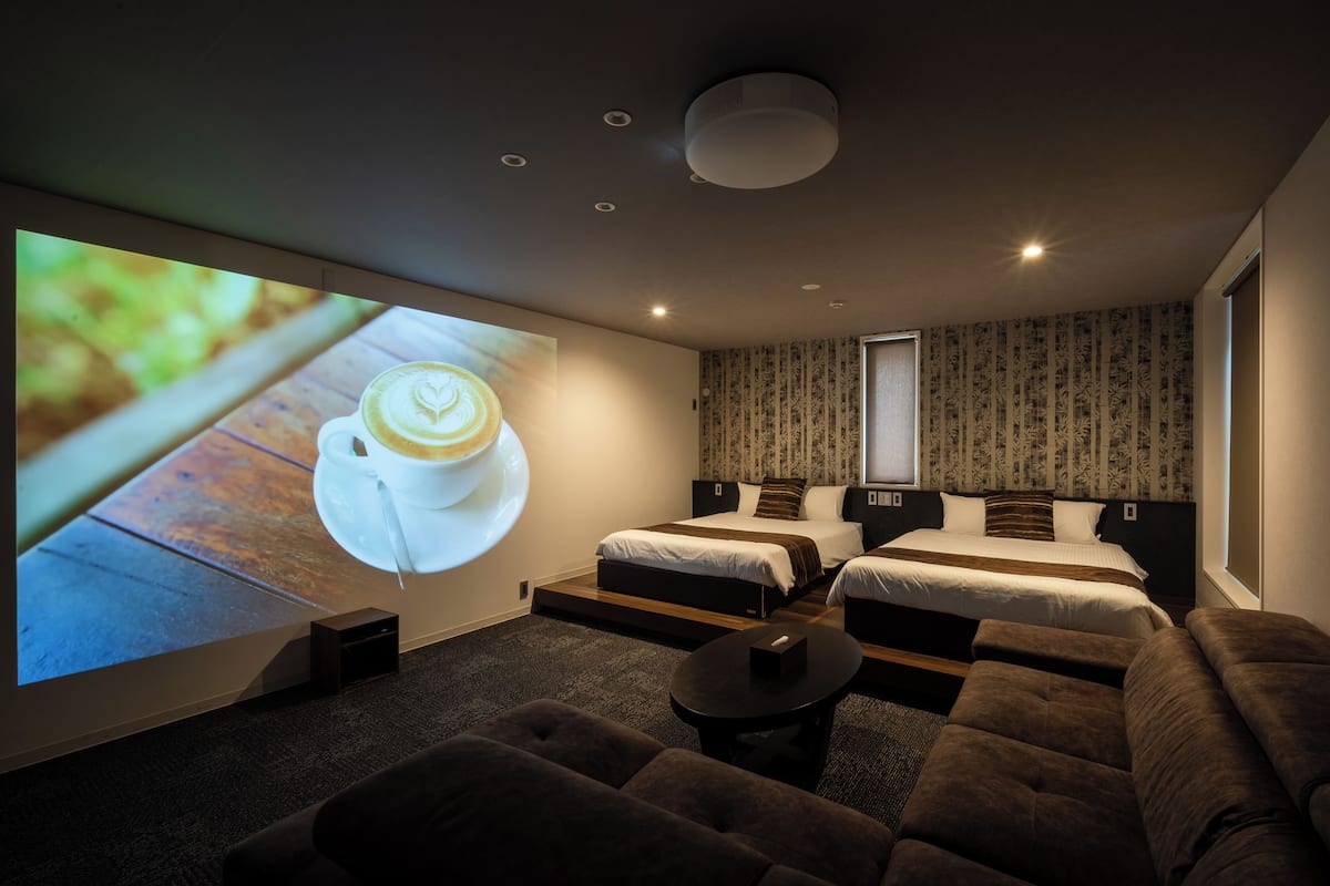 A modern hotel room with a large TV screen displaying a coffee cup, two single beds, a sofa, and a small table.