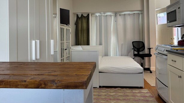 Una habitación de hotel compacta con una cama de madera, una pequeña kitchenette, un televisor y una ventana con cortinas.
