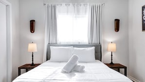 Una cama bien hecha con sábanas blancas y dos toallas enrolladas, flanqueada por dos mesitas de noche con lámparas, en una habitación con un ventanal y cortinas.