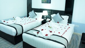 A hotel room with two beds, each adorned with white linens and red rose petals. There is a nightstand with a lamp and a phone between the beds. - Hotel Bella (Mandalay)