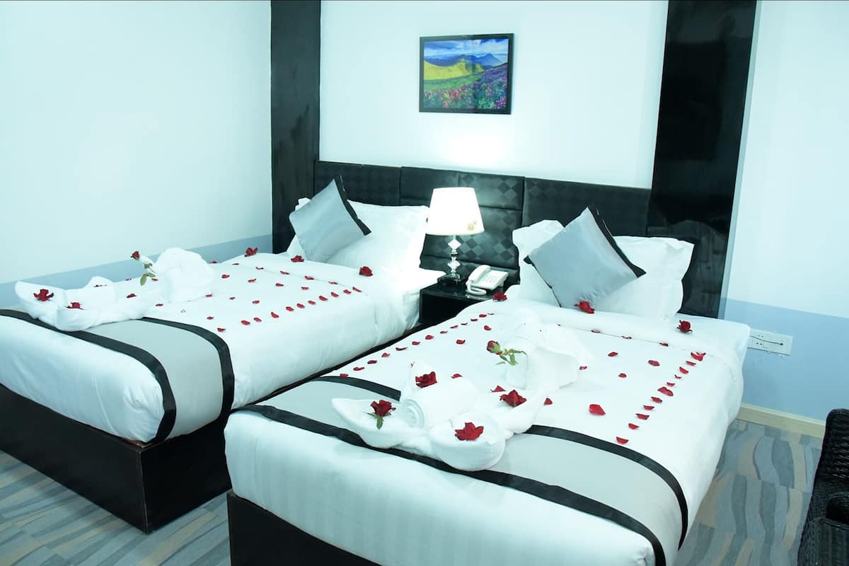 Habitación de hotel con dos camas, cada una decorada con sábanas blancas y pétalos de rosa roja. Hay una mesita de noche con una lámpara y un teléfono entre las camas.