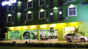 A multi-story hotel with a green facade, multiple balconies, and a parking area with motorcycles and cars. - Hotel Bella (Mandalay)
