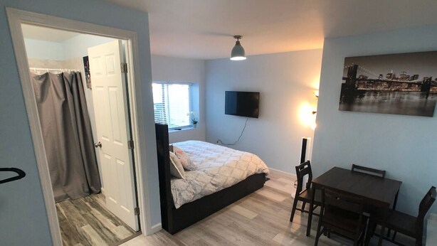 A bedroom with a bed, a desk, and a chair. There is a window with blinds, a TV mounted on the wall, and a painting on the wall. - Charming Cozy Centralize Studio (San Mateo)
