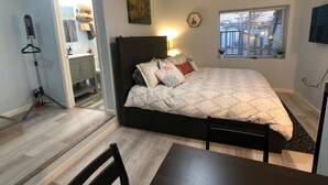 A bedroom with a bed, a table, and a chair. There is a window with blinds, a lamp, and a painting on the wall. - Charming Cozy Centralize Studio (San Mateo)
