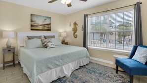 A bedroom with a bed, bedside tables, a ceiling fan, a window with blinds, and a view of a pool.