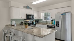A modern kitchen with white cabinets, a stainless steel refrigerator, a granite countertop, and a window with blinds.