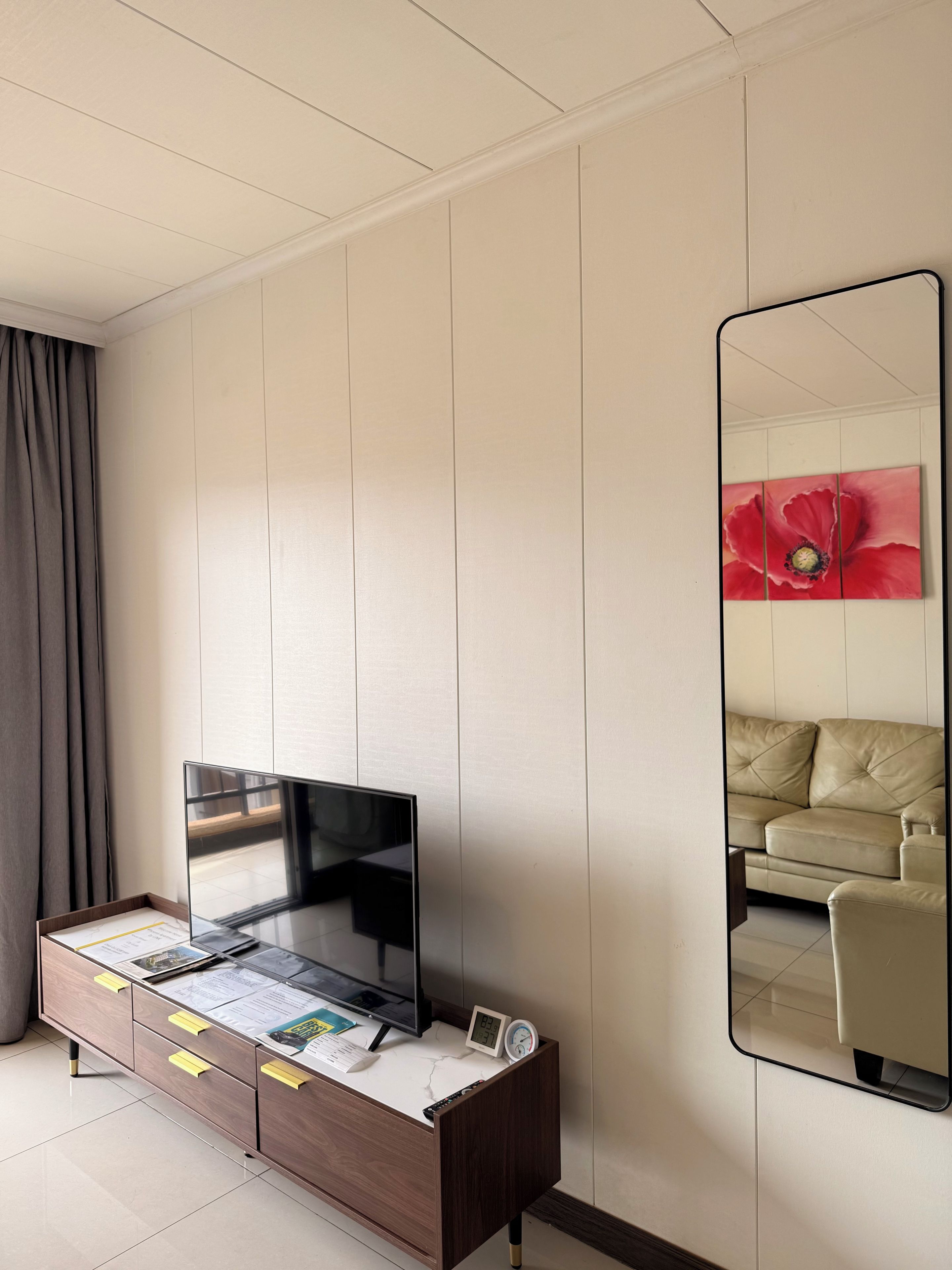 Habitación de hotel con televisor de pantalla plana sobre un mueble de madera, una mesita de noche con un reloj y un espejo grande que refleja un sofá y un cuadro de una flor roja.