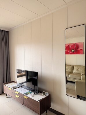 A hotel room with a flat-screen TV on a wooden cabinet, a bedside table with a clock, and a large mirror reflecting a sofa and a red flower painting. - Phoenix Apartment by LINK (Kigali)