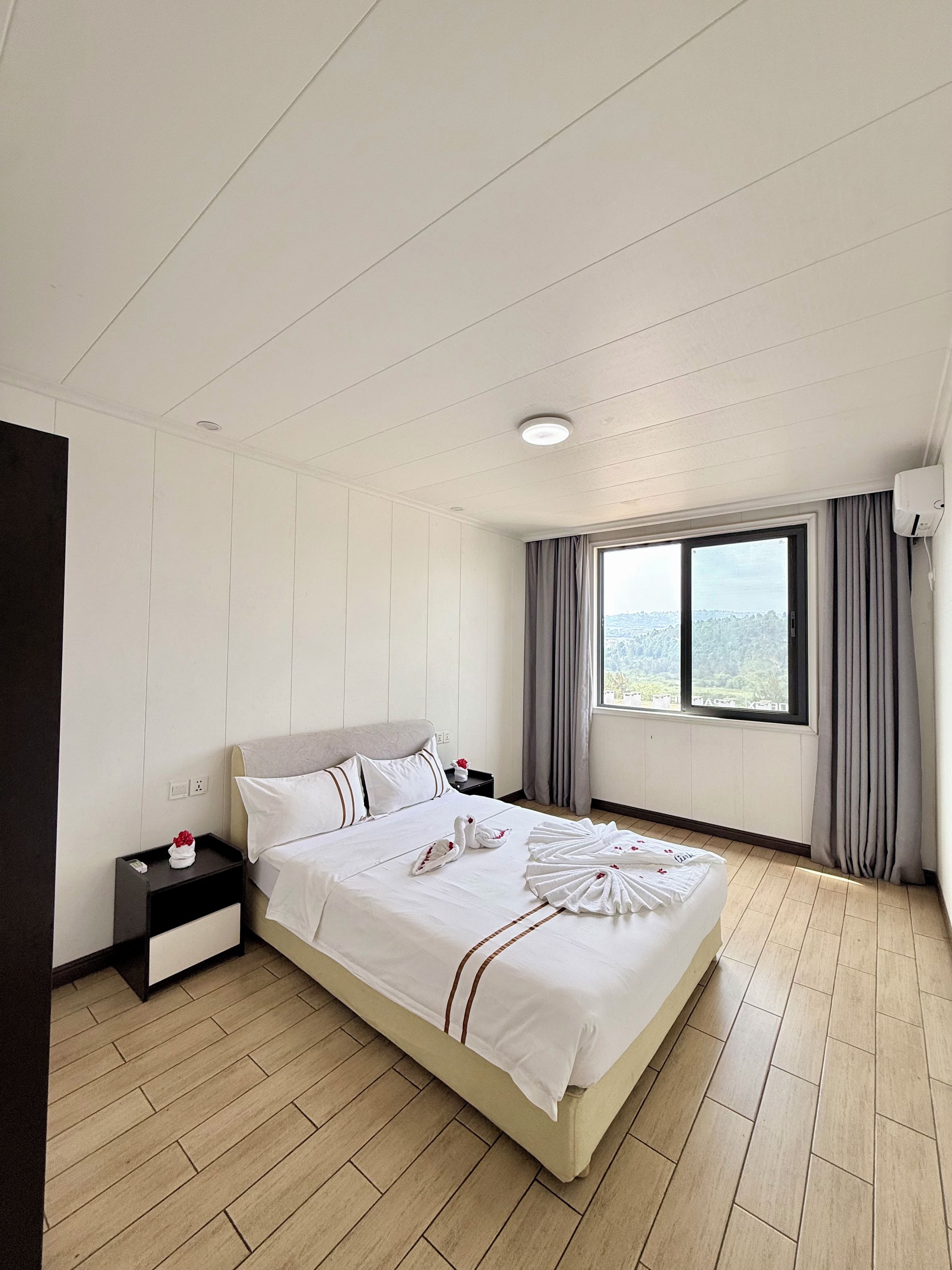 A hotel room with a large bed, a nightstand, a window with curtains, and a view of the city.