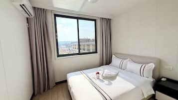 A hotel room with a bed, a window with curtains, an air conditioning unit, and a view of buildings outside.